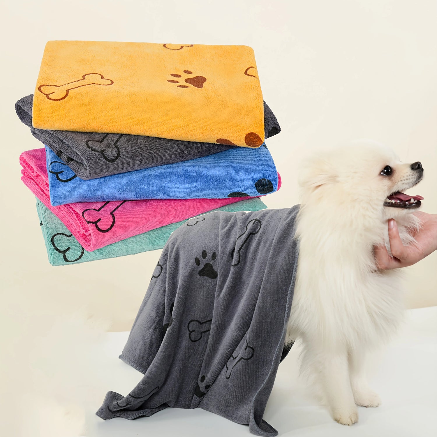 Fluffy white dog wrapped in a grey dog towel with paw and bone print, stacked colorful pet towels beside.