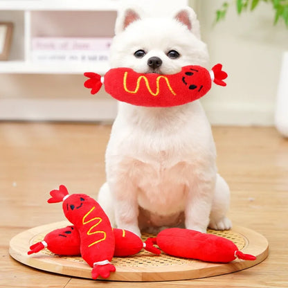White puppy holding a hot dog squeaky plush dog chew toy with red plush chew toys on a wooden mat